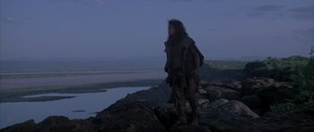 Movie still from “Quest for Fire” (1981), directed by Jean-Jacques Annaud – A man standing on top of a rock near a body of water; Wide shot, Low angle