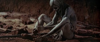 Movie still from “Quest for Fire” (1981), directed by Jean-Jacques Annaud – A person with a body paint sitting on the ground and holding a stick; Wide shot, High angle