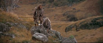 Movie still from “Quest for Fire” (1981), directed by Jean-Jacques Annaud – A man in a costume standing on top of a rock; Wide shot, Low angle