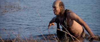Movie still from “Quest for Fire” (1981), directed by Jean-Jacques Annaud – A man with a bald head is standing in the grass near a body of water; Medium shot, Low angle