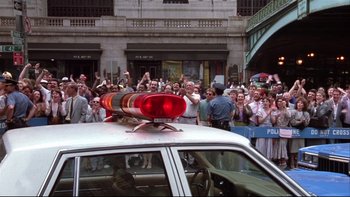 Movie still from “Quick Change” (1990), directed by Howard Franklin – A police car driving down a street with people in the background; Wide shot, High angle