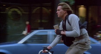 Movie still from “Quicksilver” (1986), directed by Thomas Michael Donnelly – A man riding a bike down a street with cars in the background; Medium shot, Low angle