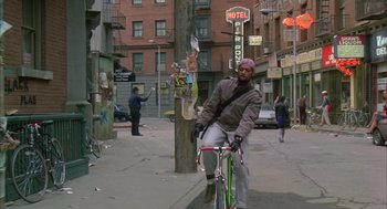 Movie still from “Quicksilver” (1986), directed by Thomas Michael Donnelly – A man riding a bike down a street next to a building; Wide shot, Low angle