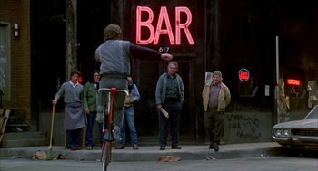 Movie still from “Quicksilver” (1986), directed by Thomas Michael Donnelly – A man riding a bike in front of a bar; Wide shot, Over the shoulder angle
