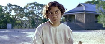Movie still from “Rabbit-Proof Fence” (2002), directed by Phillip Noyce – A young boy with brown hair is standing in front of a house; Close Up shot, High angle