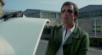 Movie still from “Rabid Dogs” (1974), directed by Mario Bava – A man wearing sunglasses and a green jacket holding a surfboard; Medium shot, Low angle