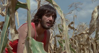 Movie still from “Rabid Dogs” (1974), directed by Mario Bava – A man with long black hair and a red tank top is in a cornfield; Close Up shot, Low angle