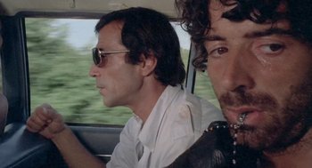 Movie still from “Rabid Dogs” (1974), directed by Mario Bava – Two men are driving in a car while wearing sunglasses; Close Up shot, Low angle