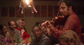 Movie still from “Rachel Getting Married” (2008), directed by Jonathan Demme – A group of people sitting at a table playing a violin; Medium shot, Over the shoulder angle