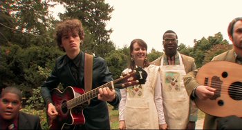 Movie still from “Rachel Getting Married” (2008), directed by Jonathan Demme – A group of people standing next to each other holding a guitar; Medium shot, Over the shoulder angle