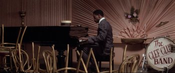Movie still from “Ragtime” (1981), directed by Milos Forman – A man sitting at a piano in front of a bunch of chairs; Medium shot, High angle