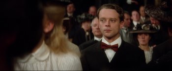 Movie still from “Ragtime” (1981), directed by Milos Forman – A man wearing a suit and a bow tie in front of a group of people; Close Up shot, Over the shoulder angle