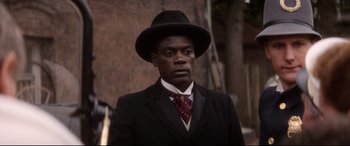 Movie still from “Ragtime” (1981), directed by Milos Forman – A man wearing a suit and a hat is standing in front of a brick wall; Close Up shot, Over the shoulder angle
