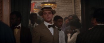 Movie still from “Ragtime” (1981), directed by Milos Forman – A man wearing a suit and a straw hat in front of a group of people; Close Up shot, Over the shoulder angle