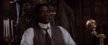 Movie still from “Ragtime” (1981), directed by Milos Forman – A man wearing a suit and tie sitting in front of a chair; Medium shot, Low angle