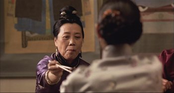 Movie still from “Raise the Red Lantern” (1991), directed by Yimou Zhang – An older woman holding a pair of chopsticks in front of a man; Close Up shot, Over the shoulder angle