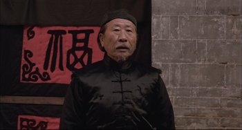 Movie still from “Raise the Red Lantern” (1991), directed by Yimou Zhang – An older man wearing a black hat and a black jacket; Medium shot, Low angle