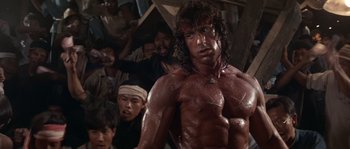 Movie still from “Rambo III” (1988), directed by Peter MacDonald – A group of people standing in a room with a shirtless man; Medium shot, Low angle