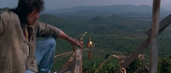 Movie still from “Rambo III” (1988), directed by Peter MacDonald – A man standing on the side of a building next to trees; Medium shot, Low angle
