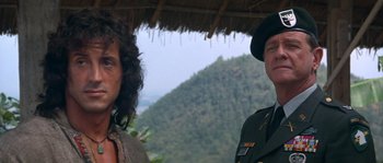 Movie still from “Rambo III” (1988), directed by Peter MacDonald – Two men standing next to each other under a thatched roof; Close Up shot, Over the shoulder angle