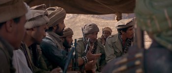 Movie still from “Rambo III” (1988), directed by Peter MacDonald – A group of men sitting around a tent talking; Medium shot, Over the shoulder angle