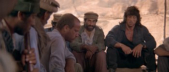 Movie still from “Rambo III” (1988), directed by Peter MacDonald – A group of men sitting next to each other in a room; Medium shot, Over the shoulder angle