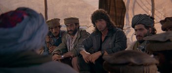 Movie still from “Rambo III” (1988), directed by Peter MacDonald – A group of men sitting next to each other on a couch; Medium shot, Over the shoulder angle