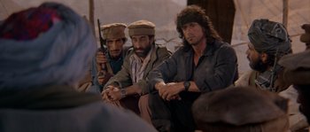 Movie still from “Rambo III” (1988), directed by Peter MacDonald – A group of men sitting next to each other on a couch; Medium shot, Over the shoulder angle
