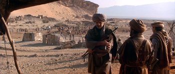 Movie still from “Rambo III” (1988), directed by Peter MacDonald – A man holding a rifle in a desert area; Wide shot, Over the shoulder angle