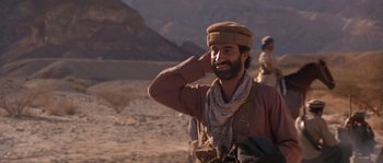 Movie still from “Rambo III” (1988), directed by Peter MacDonald – A man with a beard wearing a hat and holding his hand up to his head; Medium shot, Over the shoulder angle