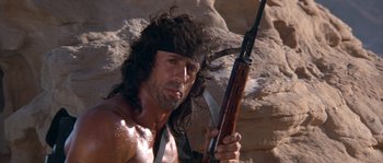 Movie still from “Rambo III” (1988), directed by Peter MacDonald – A man holding a rifle in a rocky area; Close Up shot, Low angle
