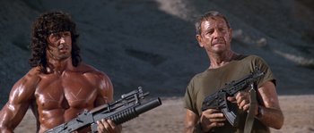 Movie still from “Rambo III” (1988), directed by Peter MacDonald – A man holding a machine gun in front of another man; Medium shot, Low angle