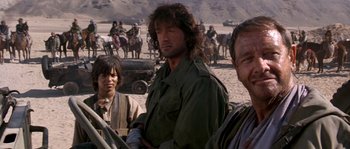 Movie still from “Rambo III” (1988), directed by Peter MacDonald – A group of men standing next to each other on top of a dirt field; Medium shot, Over the shoulder angle