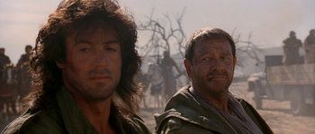 Movie still from “Rambo III” (1988), directed by Peter MacDonald – Two men standing next to each other in the desert; Close Up shot, Over the shoulder angle