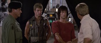Movie still from “Rambo: First Blood Part II” (1985), directed by George P. Cosmatos – A man standing next to another man on a stage; Medium shot, Low angle