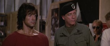Movie still from “Rambo: First Blood Part II” (1985), directed by George P. Cosmatos – A man in a military uniform standing next to another man in a red shirt; Close Up shot, Over the shoulder angle