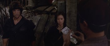 Movie still from “Rambo: First Blood Part II” (1985), directed by George P. Cosmatos – A woman sitting on the ground holding a stack of money; Medium shot, Over the shoulder angle