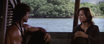 Movie still from “Rambo: First Blood Part II” (1985), directed by George P. Cosmatos – A man sitting on a dock next to a body of water; Medium shot, Over the shoulder angle