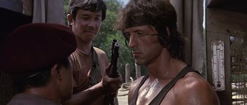 Movie still from “Rambo: First Blood Part II” (1985), directed by George P. Cosmatos – A man holding a gun while standing next to another man; Close Up shot, Over the shoulder angle