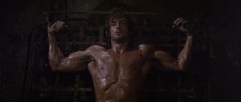 Movie still from “Rambo: First Blood Part II” (1985), directed by George P. Cosmatos – A man with a muscular body is standing in the dark; Medium shot, Low angle