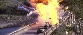 Movie still from “Rambo: First Blood Part II” (1985), directed by George P. Cosmatos – Two jeeps driving down a road with a fire coming out of it; Extreme Wide shot, High angle