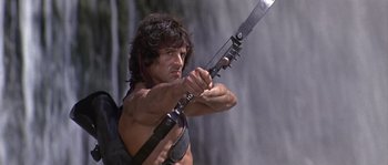 Movie still from “Rambo: First Blood Part II” (1985), directed by George P. Cosmatos – A person holding a gun; Medium shot, Low angle