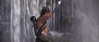 Movie still from “Rambo: First Blood Part II” (1985), directed by George P. Cosmatos – A man holding a sword in front of a waterfall; Medium shot, Low angle