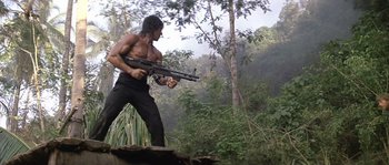 Movie still from “Rambo: First Blood Part II” (1985), directed by George P. Cosmatos – A man holding a gun while standing in the woods; Wide shot, Low angle