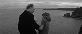 Movie still from “Rapture” (1965), directed by John Guillermin – An older man and a young woman standing next to a body of water; Medium shot, High angle