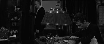 Movie still from “Rapture” (1965), directed by John Guillermin – A man standing in front of a lamp on a table; Medium shot, Over the shoulder angle