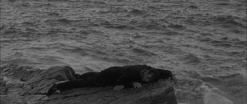 Movie still from “Rapture” (1965), directed by John Guillermin – A man laying on the ground in front of a body of water; Wide shot, High angle