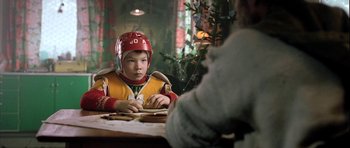 Movie still from “Rare Exports” (2010), directed by Jalmari Helander – A young boy wearing a football helmet while sitting at a table; Medium shot, Over the shoulder angle