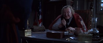 Movie still from “Ravenous” (1999), directed by Antonia Bird – An older man sitting at a table with a flag in the background; Medium shot, High angle