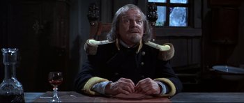 Movie still from “Ravenous” (1999), directed by Antonia Bird – An older man wearing a uniform sitting at a table; Medium shot, High angle